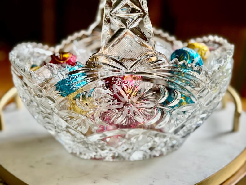 May include: A clear, cut-glass basket with a handle, filled with colorful foil-wrapped candies. The basket has a scalloped rim and a floral design on the base. The candies are in shades of red, blue, and yellow.