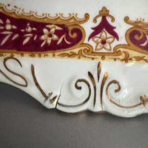May include: A white porcelain plate with a gold and burgundy floral design. The plate has a scalloped edge and a small chip on the rim.