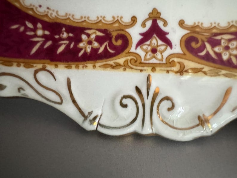 May include: A white porcelain plate with a gold and burgundy floral design. The plate has a scalloped edge and a small chip on the rim.