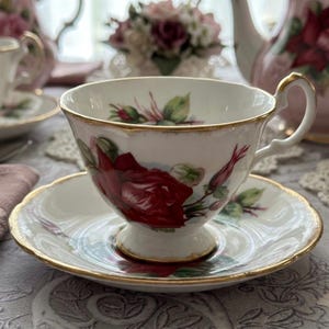 May include: White teacup and saucer set with a red rose floral design and gold trim. The teacup has a curved handle and sits on a matching saucer. A teapot and other teacups are visible in the background.