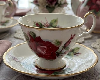May include: White teacup and saucer set with a red rose floral design and gold trim. The teacup has a curved handle and sits on a matching saucer. A teapot and other teacups are visible in the background.
