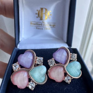 Op de afbeelding: Een paar hartvormige oorbellen in een blauwe doos. Elke oorbel heeft drie pastelkleurige harten in roze, lila en turquoise, met heldere kristallen en een goudkleurig montuur. De doos is van Bradford Exchange.