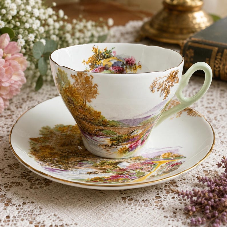 May include: A white porcelain teacup and saucer set featuring a colorful landscape illustration of a stone bridge over a river, surrounded by trees and flowers. The teacup has a scalloped rim and a pale green handle, with gold trim on the edges.