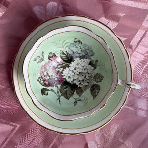 May include: A pale green porcelain teacup and saucer set featuring a scalloped edge with gold trim. The interior of the cup and the center of the saucer are decorated with a detailed floral illustration of white, pink, and blue hydrangea blossoms.