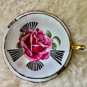May include: A white teacup and saucer set with a gold rim and handle. The saucer features a large pink rose with green leaves and black and white fan-shaped designs. The teacup is on a white, fluffy surface.