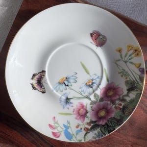 Może przedstawiać: Biały spodek porcelanowy z pozłacanym rantem przedstawiający wz&oacute;r kwiatowy z motylami. Kwiaty są r&oacute;żowe, białe, niebieskie i ż&oacute;łte. Motyle są brązowe, r&oacute;żowe i fioletowe.