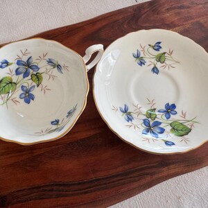 May include: A white teacup and saucer set with a blue and green floral pattern. The floral design features delicate blue flowers and green leaves.