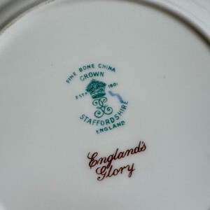 May include: Close-up of a white fine bone china plate with a green and brown emblem. The emblem includes a crown, the words "Fine Bone China Crown", and "England's Glory".