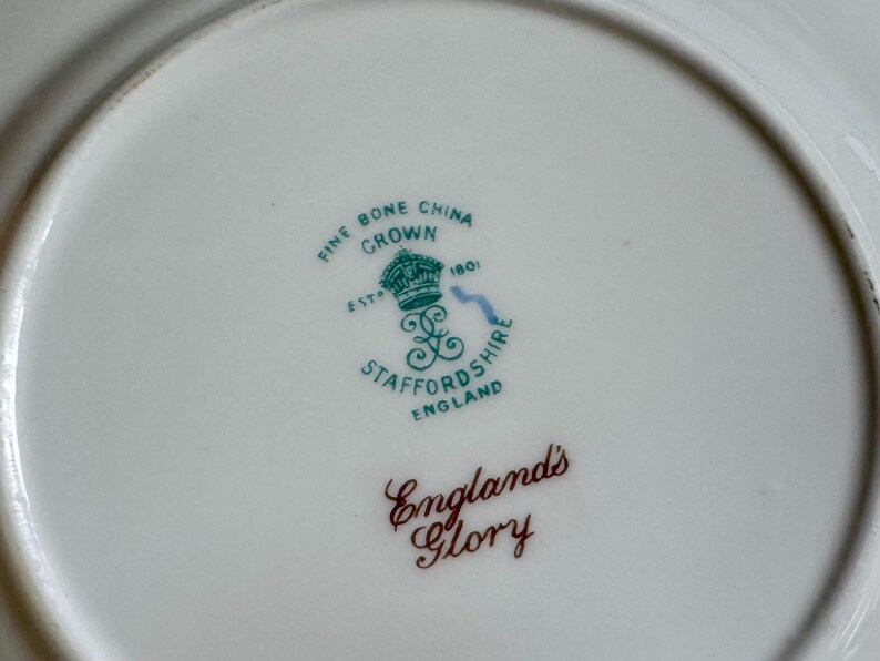 May include: Close-up of a white fine bone china plate with a green and brown emblem. The emblem includes a crown, the words "Fine Bone China Crown", and "England's Glory".
