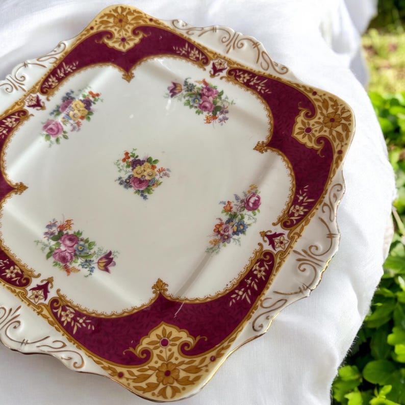 May include: A square, white ceramic serving plate with a burgundy and gold border. The plate has a scalloped edge and is decorated with floral designs.