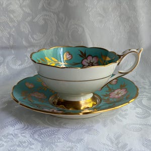 May include: A porcelain teacup and saucer set featuring a vibrant turquoise interior and rim with delicate pink floral motifs and gold leaf accents. The cup has a scalloped edge, a pedestal base, and a curved handle, all trimmed in polished gold.