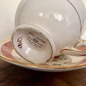 Pode incluir: Conjunto de ch&aacute; Royal Stafford em porcelana branca. O pires tem um padr&atilde;o floral vermelho e dourado. A ch&aacute;vena e o pires t&ecirc;m acabamentos dourados. O texto "Royal Stafford Bone China Made in England Est 1845" &eacute; vis&iacute;vel.