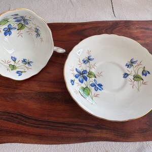 May include: A white teacup and saucer set with a blue and green floral pattern. The floral design features delicate blue flowers and green leaves.