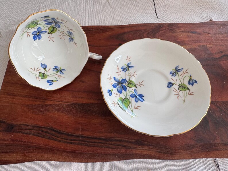 May include: A white teacup and saucer set with a blue and green floral pattern. The floral design features delicate blue flowers and green leaves.