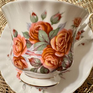 Pode incluir: Um conjunto de ch&aacute;vena e pires branco com um design floral com rosas laranja e rosa. A ch&aacute;vena de ch&aacute; tem uma borda dourada e o pires tem uma borda ondulada.
