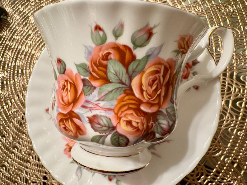 Pode incluir: Um conjunto de ch&aacute;vena e pires branco com um design floral com rosas laranja e rosa. A ch&aacute;vena de ch&aacute; tem uma borda dourada e o pires tem uma borda ondulada.