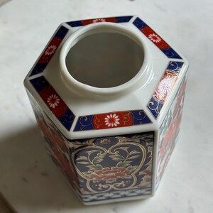 Peut inclure: Un vase hexagonal en c&eacute;ramique blanche avec un motif floral. Le vase pr&eacute;sente des accents rouges, bleus et dor&eacute;s. Le motif floral comprend des fleurs rouges et des feuilles vertes. Le vase a une large ouverture.