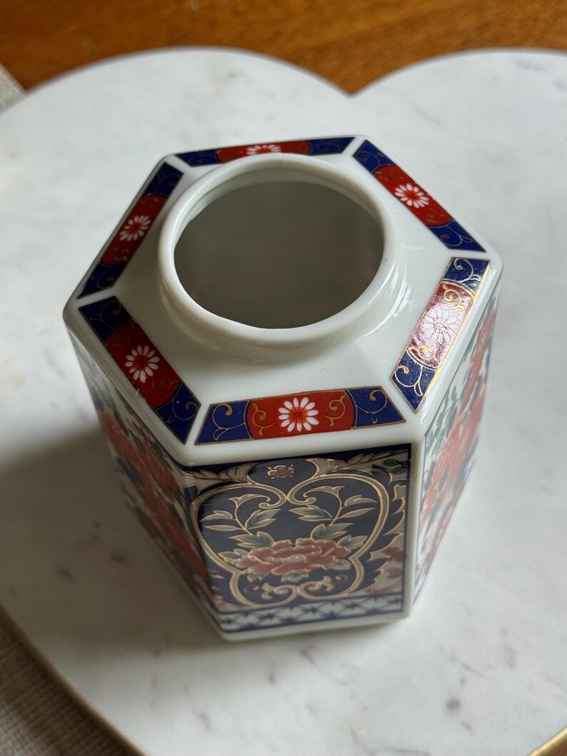 Peut inclure: Un vase hexagonal en c&eacute;ramique blanche avec un motif floral. Le vase pr&eacute;sente des accents rouges, bleus et dor&eacute;s. Le motif floral comprend des fleurs rouges et des feuilles vertes. Le vase a une large ouverture.