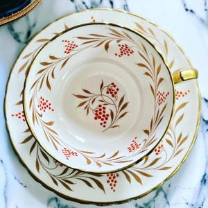 Pu&ograve; includere: Un set di tazza e piattino da t&egrave; vintage. La tazza e il piattino in porcellana bianca sono adornati con finiture dorate e un motivo di foglie dorate e bacche rosse. La tazza ha un manico dorato e il piattino ha un bordo smerlato.