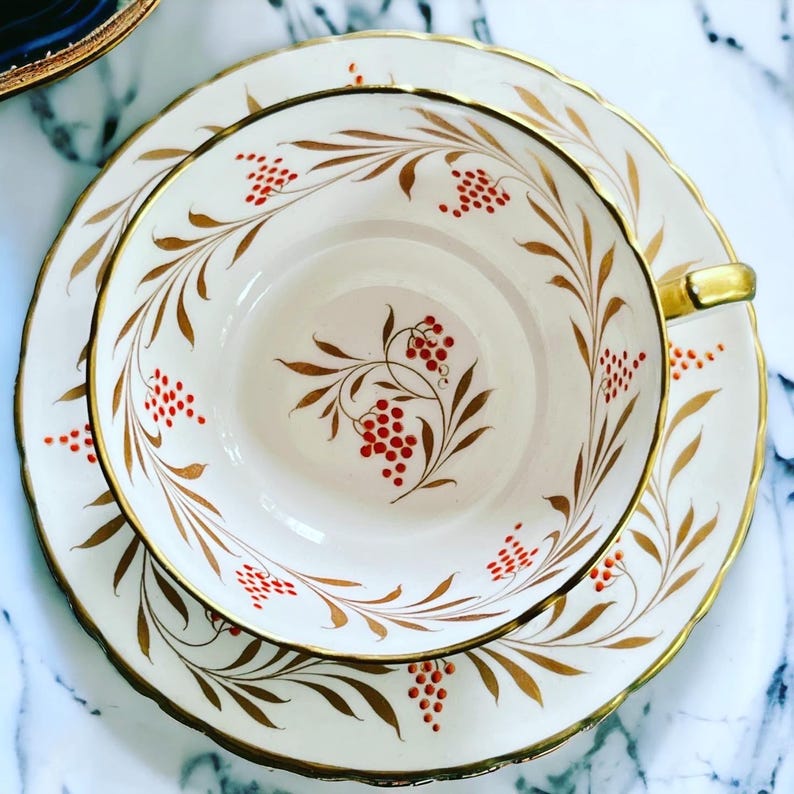 Pu&ograve; includere: Un set di tazza e piattino da t&egrave; vintage. La tazza e il piattino in porcellana bianca sono adornati con finiture dorate e un motivo di foglie dorate e bacche rosse. La tazza ha un manico dorato e il piattino ha un bordo smerlato.