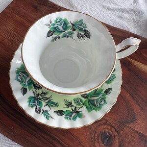 May include: A white teacup and saucer with a green and white floral pattern. The cup has a gold rim and the saucer has a scalloped edge.
