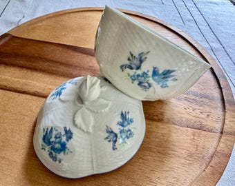 May include: A white ceramic trinket box with a lid, featuring a textured surface and blue bird and floral designs. The lid is topped with a white rose bud. The box is on a wooden tray.