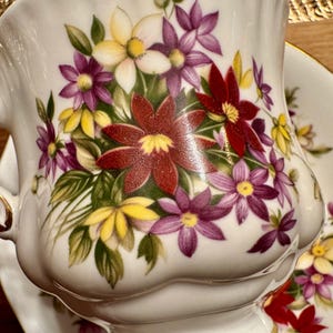 May include: White porcelain teacup and saucer with a floral design. The cup and saucer feature a bouquet of red, purple, yellow, and white flowers with green leaves.