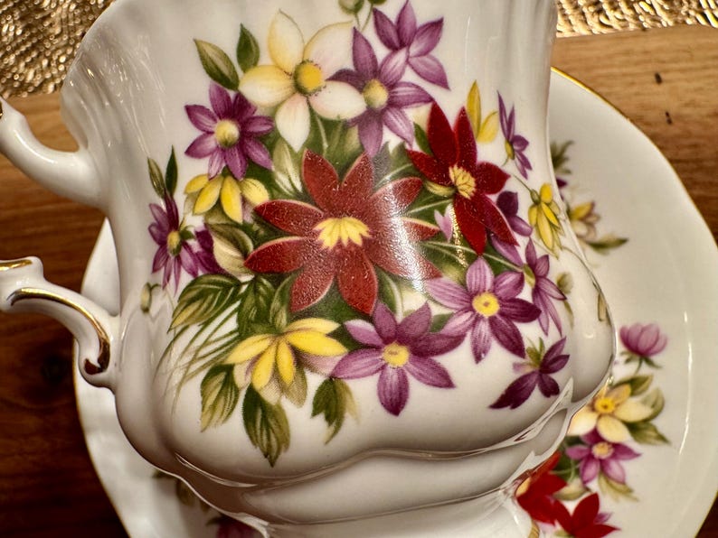 May include: White porcelain teacup and saucer with a floral design. The cup and saucer feature a bouquet of red, purple, yellow, and white flowers with green leaves.