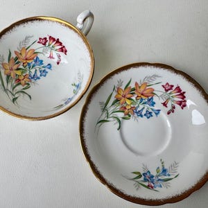 May include: A white teacup and saucer set with a floral design. The cup and saucer have a gold rim and feature a delicate pattern of yellow, orange, red, and blue flowers.