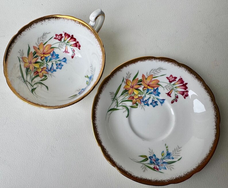 May include: A white teacup and saucer set with a floral design. The cup and saucer have a gold rim and feature a delicate pattern of yellow, orange, red, and blue flowers.