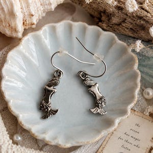 K&ouml;nnte beinhalten: Silberne Meerjungfrau-Ohrringe auf einer hellblauen, wellenf&ouml;rmigen Schale. Die Ohrringe haben ein detailliertes Meerjungfrau-Design. Die Schale ist von Muscheln, Perlen und einer kleinen Karte mit dem Text "the ocean has magic for those who listen" umgeben.