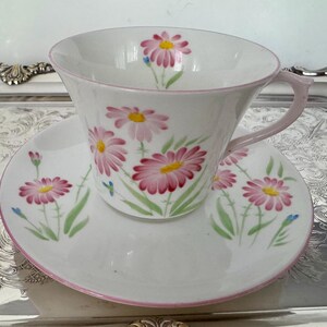 May include: White teacup and saucer set with a pink daisy floral pattern. The cup and saucer have a pink trim and handle. The daisies have pink petals and yellow centers, with green stems and leaves.