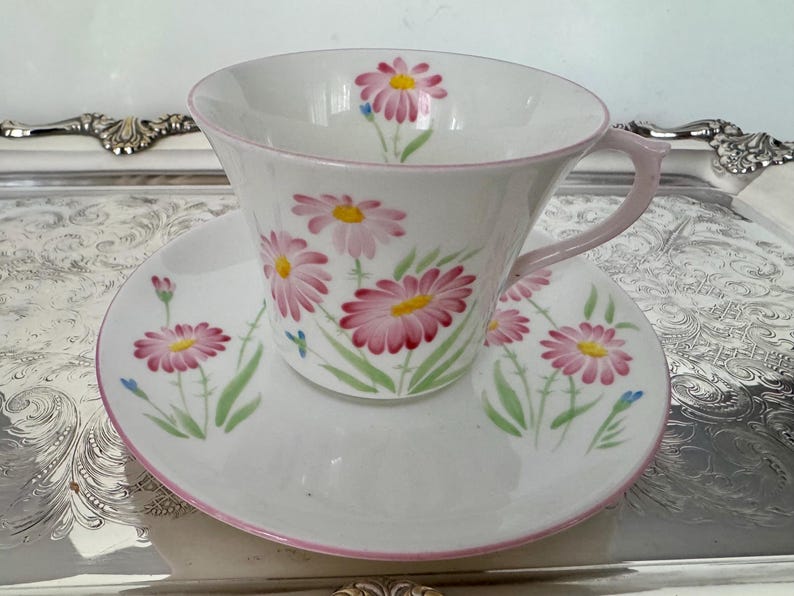May include: White teacup and saucer set with a pink daisy floral pattern. The cup and saucer have a pink trim and handle. The daisies have pink petals and yellow centers, with green stems and leaves.