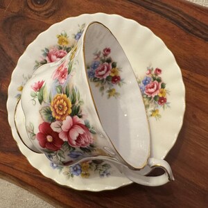 May include: A white teacup and saucer with a floral pattern in shades of pink, red, yellow, and blue. The cup is tilted and the saucer has a scalloped edge.