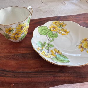 May include: A white teacup and saucer with a floral pattern of yellow flowers and green leaves. The cup has a scalloped edge and a gold trim.