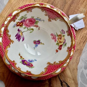 May include: A vintage teacup and saucer set with a floral design. The cup and saucer feature a white background with pink, gold, and floral accents. The rim of the saucer has a pink scale pattern. The cup has a white handle.