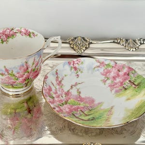 May include: A vintage teacup and saucer set. The white porcelain cup and saucer are decorated with pink floral patterns and gold trim. The saucer has a scalloped edge. The set is displayed on a silver tray.