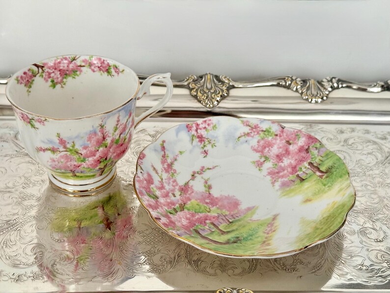 May include: A vintage teacup and saucer set. The white porcelain cup and saucer are decorated with pink floral patterns and gold trim. The saucer has a scalloped edge. The set is displayed on a silver tray.