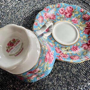 K&ouml;nnte beinhalten: Ein blaues und wei&szlig;es Tee-Set mit Blumenmuster. Die Untertasse hat einen goldenen Rand. Die Teetasse hat ein Royal Albert Logo auf der Unterseite, das "Bone China, Merrie England Series, Harewood" lautet.