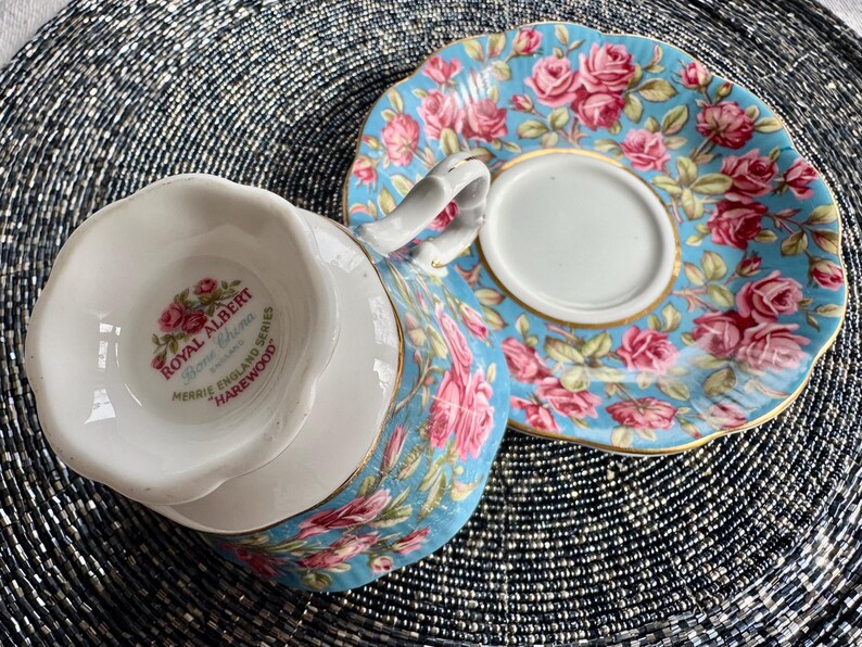 K&ouml;nnte beinhalten: Ein blaues und wei&szlig;es Tee-Set mit Blumenmuster. Die Untertasse hat einen goldenen Rand. Die Teetasse hat ein Royal Albert Logo auf der Unterseite, das "Bone China, Merrie England Series, Harewood" lautet.