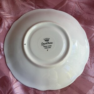 May include: A white bone china saucer with a scalloped edge. The underside features a black stamp with a crown icon and the text Queen Anne, Bone China, England. The saucer has a circular foot rim and a smooth, glossy finish on the surface.