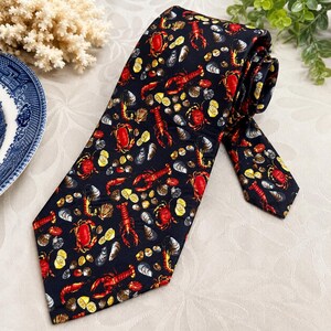 Puede incluir: Una corbata negra con un patr&oacute;n repetitivo de langostas rojas, cangrejos rojos, conchas marinas grises y rodajas de lim&oacute;n amarillas. La corbata tiene una punta afilada y una estructura plegada, mostrando un estampado vibrante de mariscos.