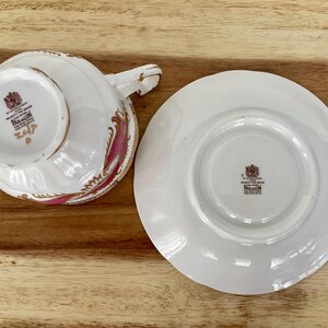 May include: White teacup and saucer set with a pink and gold floral design. The teacup has a handle and the saucer is round. Both pieces have a crest and the word "PARAGON" printed on them. The set is on a wooden surface.