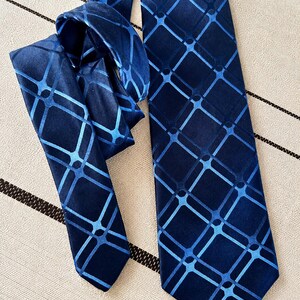 May include: A navy blue tie with a geometric pattern of light blue squares and lines. The tie is knotted, with the wider end and the knot visible. The pattern is repeated across the entire tie, creating a cohesive design.