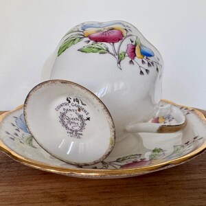May include: A vintage white teacup and saucer set with a floral pansy design. The cup and saucer have gold trim. The saucer has a small spoon rest. The teacup is upside down, revealing the bottom of the cup and saucer.