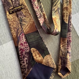 Pu&ograve; includere: Una cravatta con una stampa paesaggistica multicolore con alberi ed edifici nei toni del marrone, verde e crema. La cravatta ha un'etichetta nera cucita sul retro con la scritta St. Stefano 100% Silk Italy. La fodera &egrave; in tessuto blu scuro.