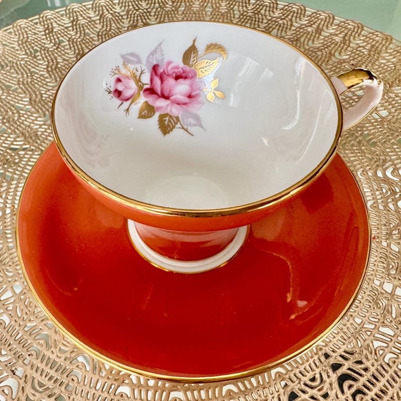 May include: A vintage teacup and saucer set with a floral design. The cup is white with a gold rim and a pink rose with gold accents. The saucer is a bright orange color with a gold rim.