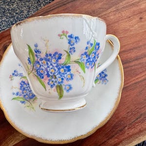 May include: A white teacup and saucer with a floral pattern of blue forget-me-nots and pink flowers. The edges of the saucer and cup are trimmed with gold.