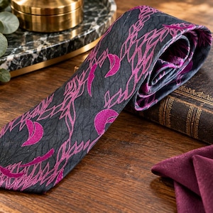 Puede incluir: Una corbata con una base de color gris carb&oacute;n oscuro y un patr&oacute;n repetitivo de lunas crecientes de color magenta vibrante y formas estilizadas similares a hojas. La tela tiene una textura sutil y la corbata est&aacute; doblada para mostrar la punta.