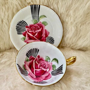 May include: A vintage teacup and saucer set. The white porcelain cup and saucer feature a large pink rose design with green leaves, black and white striped accents, and gold trim. The cup has a gold handle.